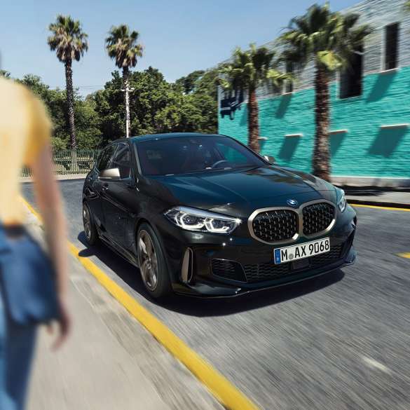 BMW M135i xDrive, trojštvrťový predný pohľad. BMW M135i xDrive v metalickej farbe Black Sapphire, trojštvrťový predný pohľad počas jazdy, so ženou v popredí.