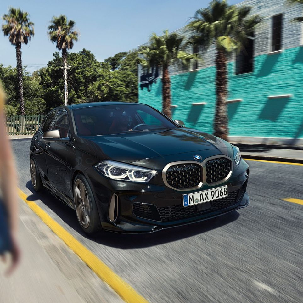 BMW M135i xDrive, trojštvrťový predný pohľad. BMW M135i xDrive v metalickej farbe Black Sapphire, trojštvrťový predný pohľad počas jazdy, so ženou v popredí.