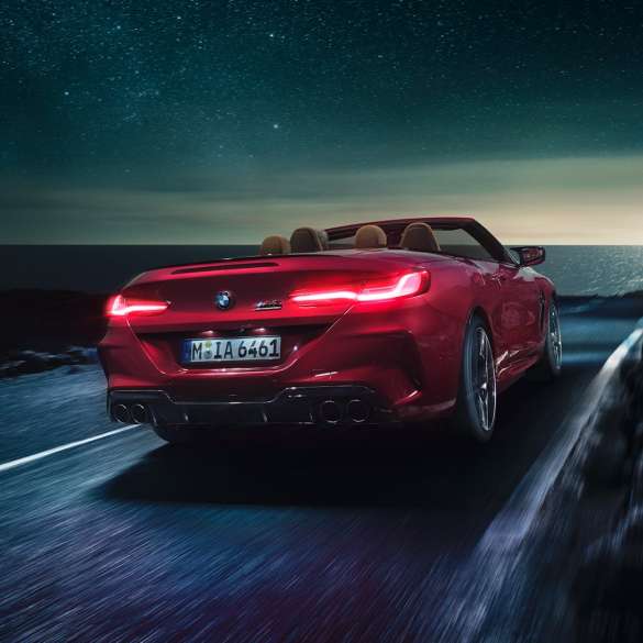 BMW M8 Competition Cabrio, trojštvrťový zadný pohľad. BMW M8 Competition Cabrio v metalickej farbe Motegi Red s otvorenou strechou, trojštvrťový zadný pohľad, vozidlo jazdiace v noci po pobrežnej ceste.