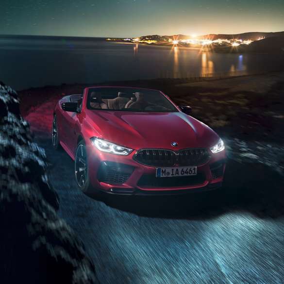 BMW M8 Competition Cabrio, trojštvrťový predný pohľad. BMW M8 Competition Cabrio v metalickej farbe Motegi Red s otvorenou strechou, trojštvrťový bočný pohľad, vozidlo jazdiace v noci po pobrežnej ceste.