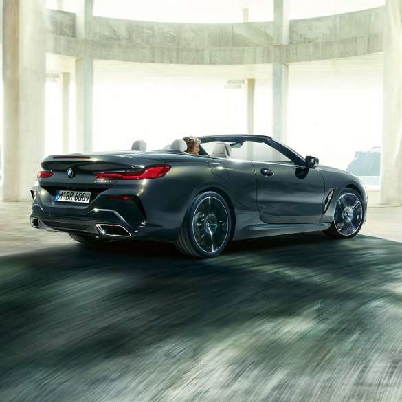 BMW M850i xDrive Cabrio, trojštvrťový zadný pohľad. BMW M850i xDrive Cabrio v metalickej farbe Dravit Grey s otvorenou strechou, trojštvrťový zadný pohľad, vozidlo jazdiace cez betónovú budovu.