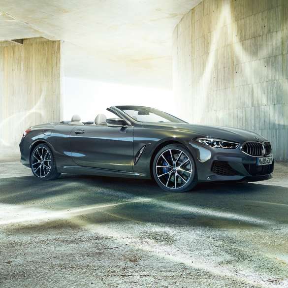 BMW M850i xDrive Cabrio, trojštvrťový bočný pohľad. BMW M850i xDrive Cabrio v metalickej farbe Dravit Grey s otvorenou strechou, trojštvrťový bočný pohľad, vozidlo stojace pred betónovou budovou.