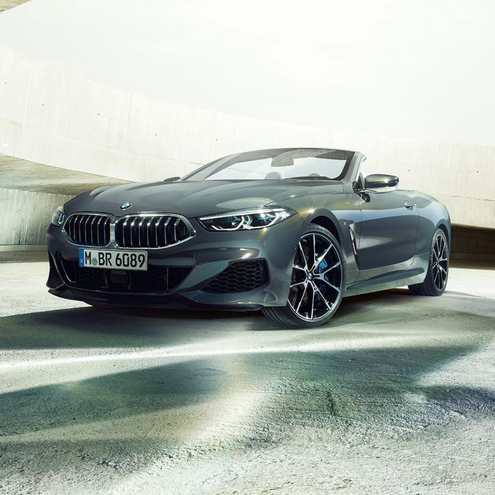 BMW M850i xDrive Cabrio, trojštvrťový predný pohľad. BMW M850i xDrive Cabrio v metalickej farbe Dravit Grey s otvorenou strechou, trojštvrťový predný pohľad, vozidlo stojace v betónovej budove.