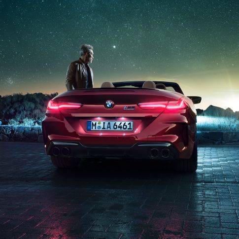 BMW M8 Competition Cabrio, zadný pohľad. BMW M8 Competition Cabrio v metalickej farbe Motegi Red s otvorenou strechou, zadný pohľad, stojace vozidlo s mužom pod hviezdnou oblohou.