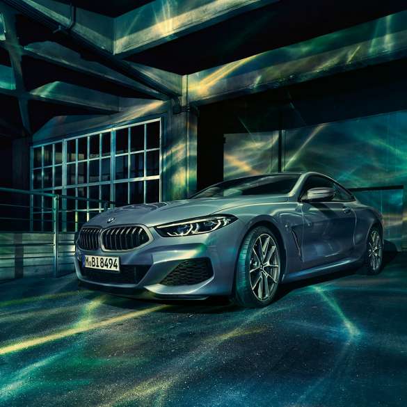 BMW M850i xDrive Coupé, trojštvrťový predný pohľad. BMW M850i xDrive Coupé v metalickej farbe Barcelona Blue, trojštvrťový predný pohľad pred priečelím budovy.
