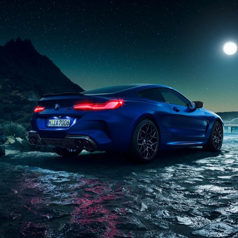 BMW M8 Competition Coupé, trojštvrťový zadný pohľad. BMW M8 Competition Coupé v metalickej farbe Marina Bay Blue s rozsvietenými LED zadnými svetlami, trojštvrťový statický zadný pohľad počas splnu mesiaca.