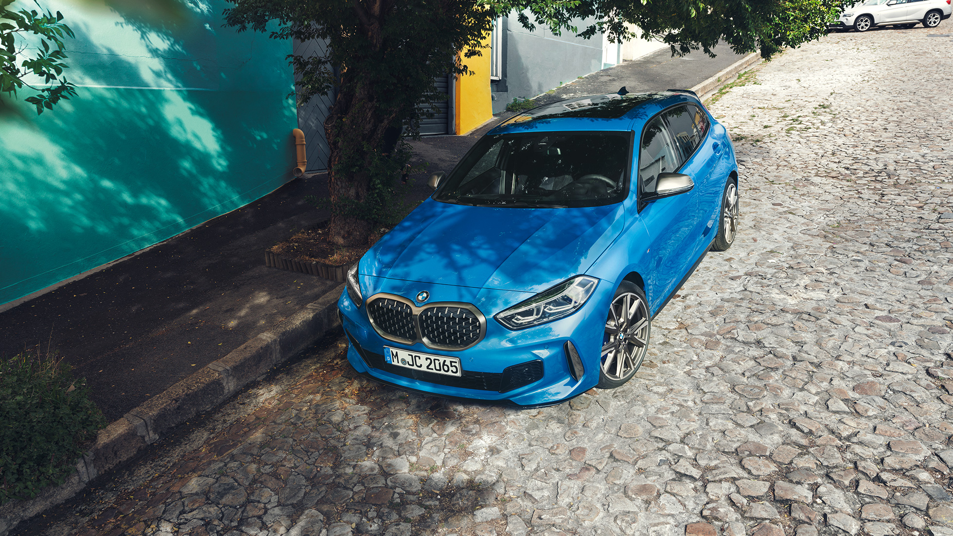 BMW radu 1 odstavené pri ceste. BMW radu 1 v metalickej farbe Misano Blue, trojštvrťový predný pohľad, parkovanie pri ceste z vtáčej perspektívy.