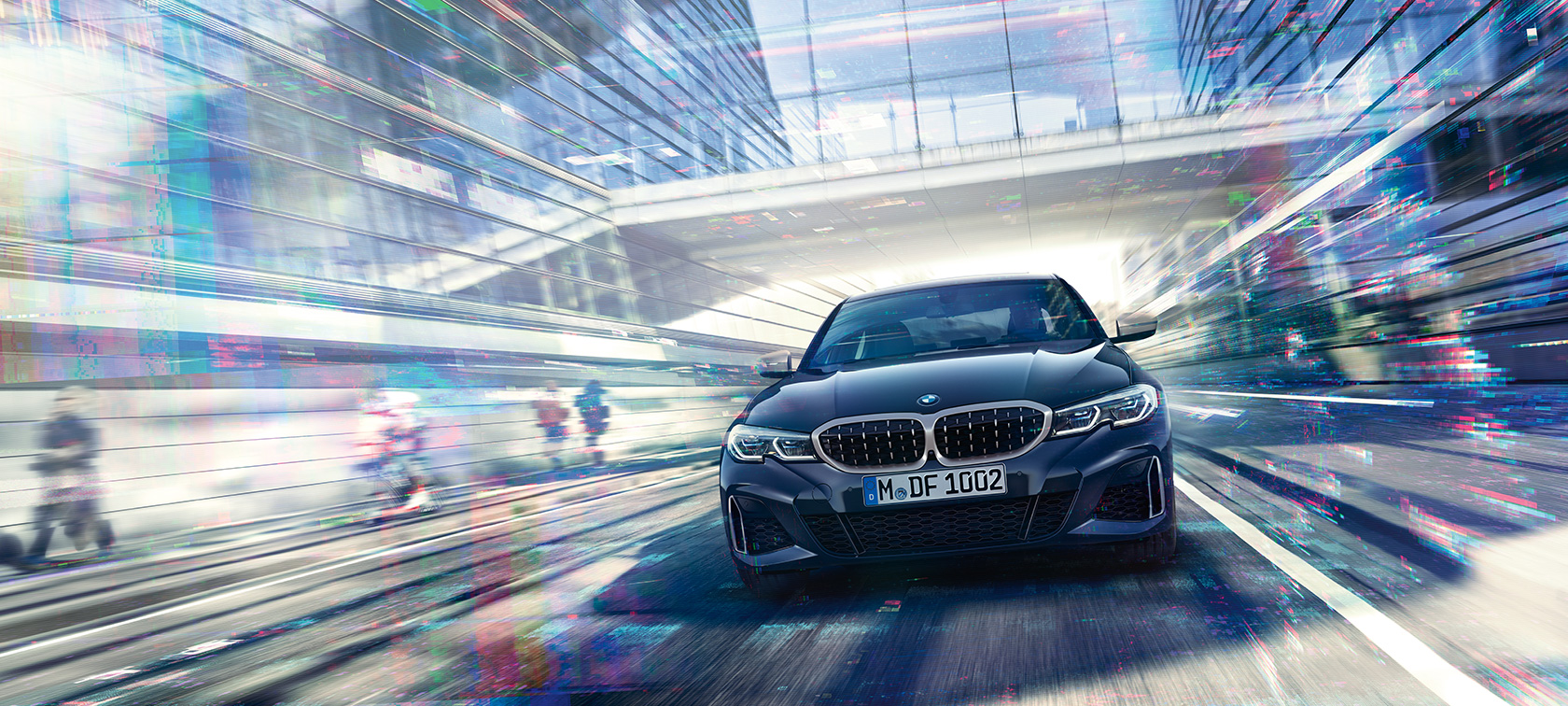 BMW radu 3 Sedan, čelný pohľad na BMW M340i xDrive. Čelný jazdný pohľad na BMW M340i xDrive Sedan medzi sklenenými mestskými budovami.
