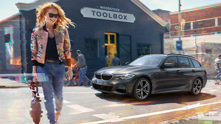 BMW radu 3 Touring (G21), predobočný záber so ženou pred drevárskou dielňou. Žena prechádza cez cestu pred BMW radu 3 Touring (G21) pred drevárskou dielňou.