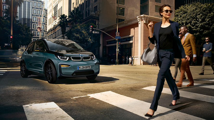BMW i3 I01 predobočný pohľad so ženou kráčajúcou cez prechod BMW i3 I01 Blue Ridge Mountain so zvýraznením v metalíze Frozen Grey predobočný pohľad so ženou kráčajúcou cez prechod