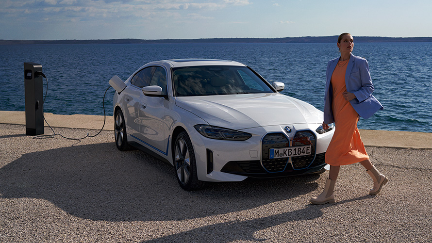Predobočný záber BMW i4 eDrive40 G26 2021 na nabíjacej stanici Žena pred BMW i4 eDrive40 G26 2021 Mineral White predobočný záber na nabíjacej stanici pred jazerom