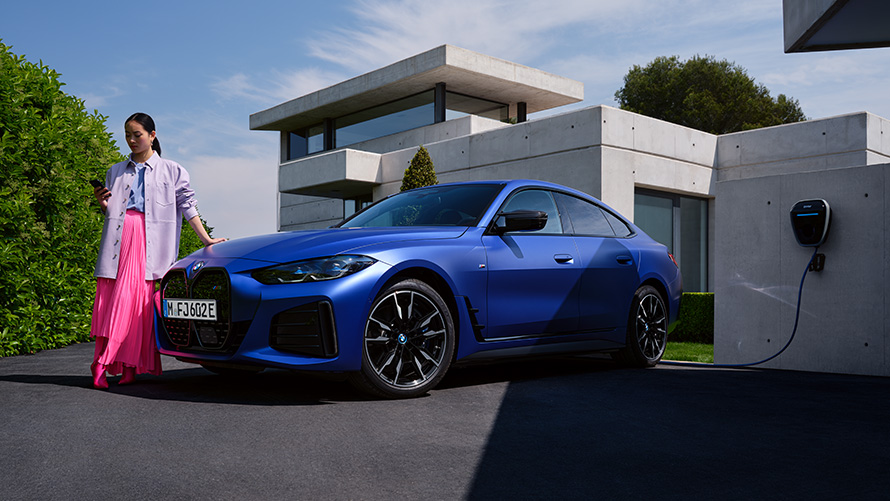Bočný záber BMW i4 M50 G26 2021 so ženou pred domom BMW i4 M50 G26 2021 BMW Individual metalíza Frozen Portimao Blue bočný pohľad so ženou pred domom