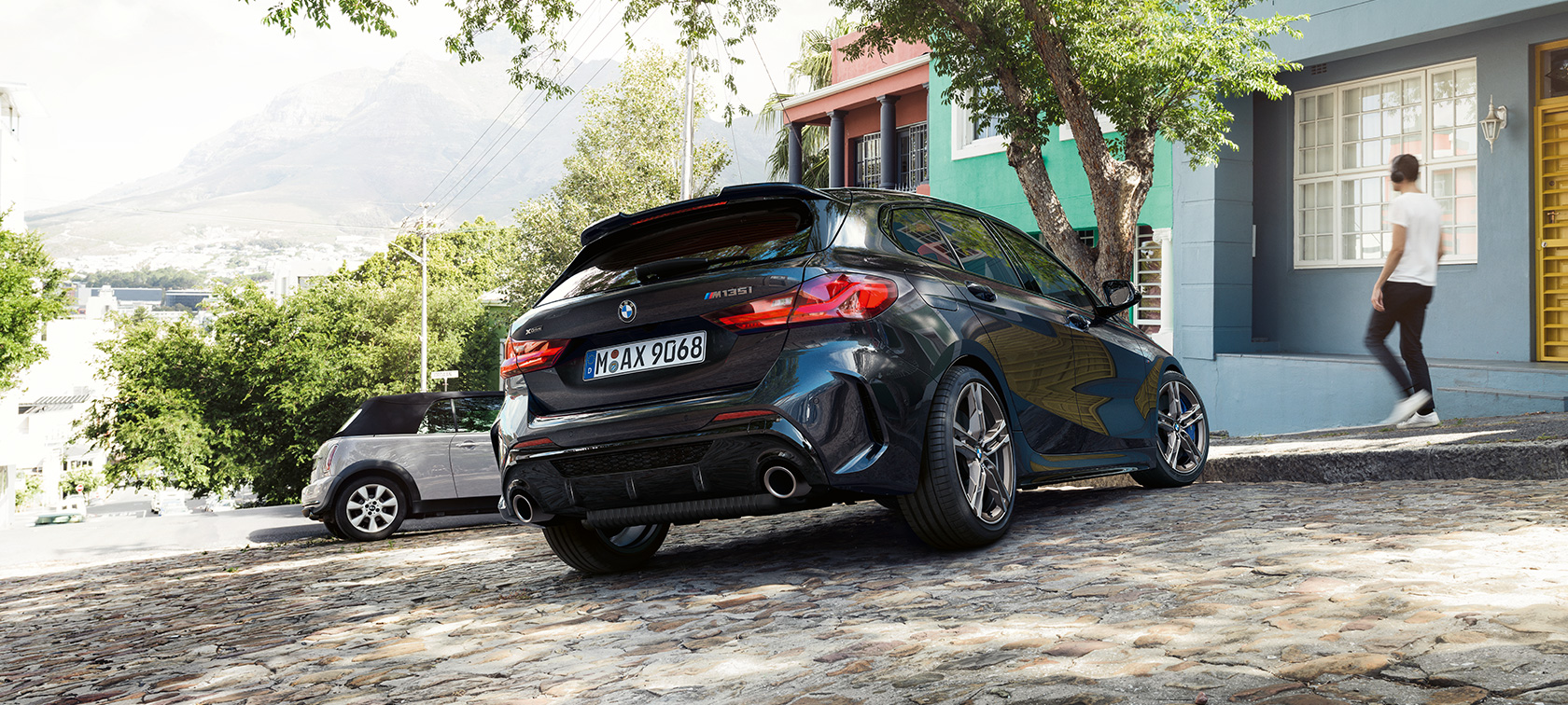 BMW M135i xDrive, trojštvrťový zadný pohľad. BMW M135i xDrive v metalickej farbe Black Sapphire, trojštvrťový zadný pohľad na odparkované vozidlo pred rezidenčnou budovou.