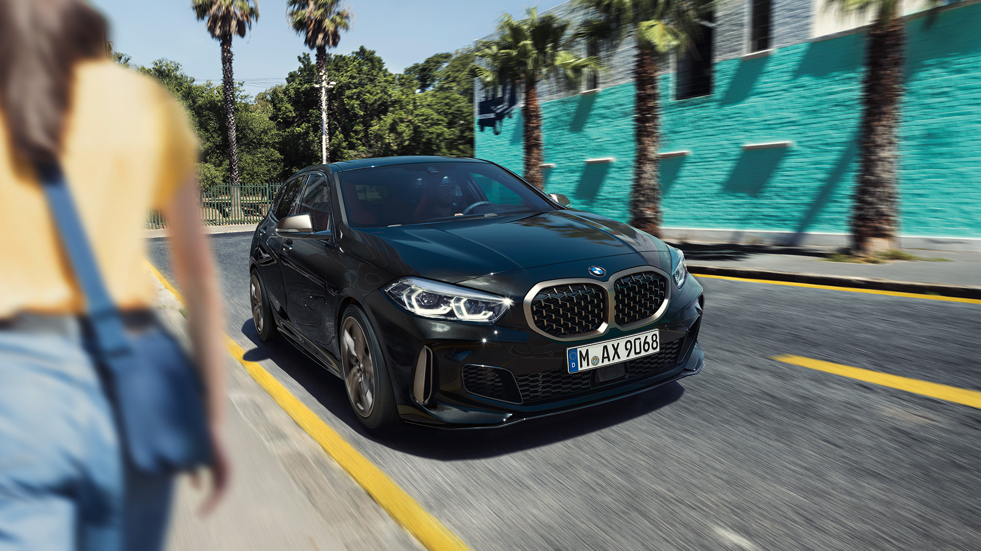 BMW M135i xDrive, trojštvrťový predný pohľad. BMW M135i xDrive v metalickej farbe Black Sapphire, trojštvrťový predný pohľad počas jazdy, so ženou v popredí.