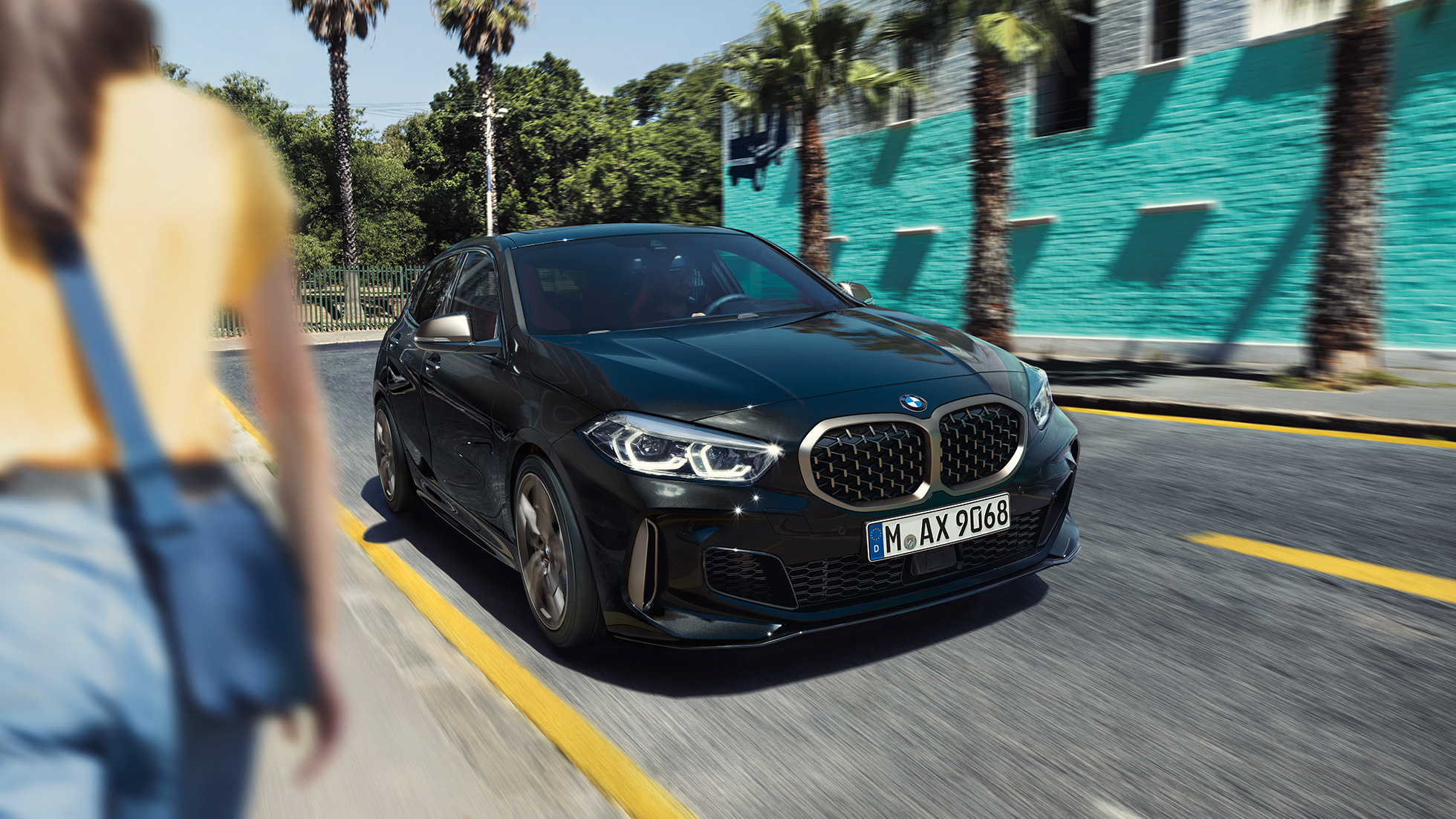 BMW M135i xDrive, trojštvrťový predný pohľad. BMW M135i xDrive v metalickej farbe Black Sapphire, trojštvrťový predný pohľad počas jazdy, so ženou v popredí.