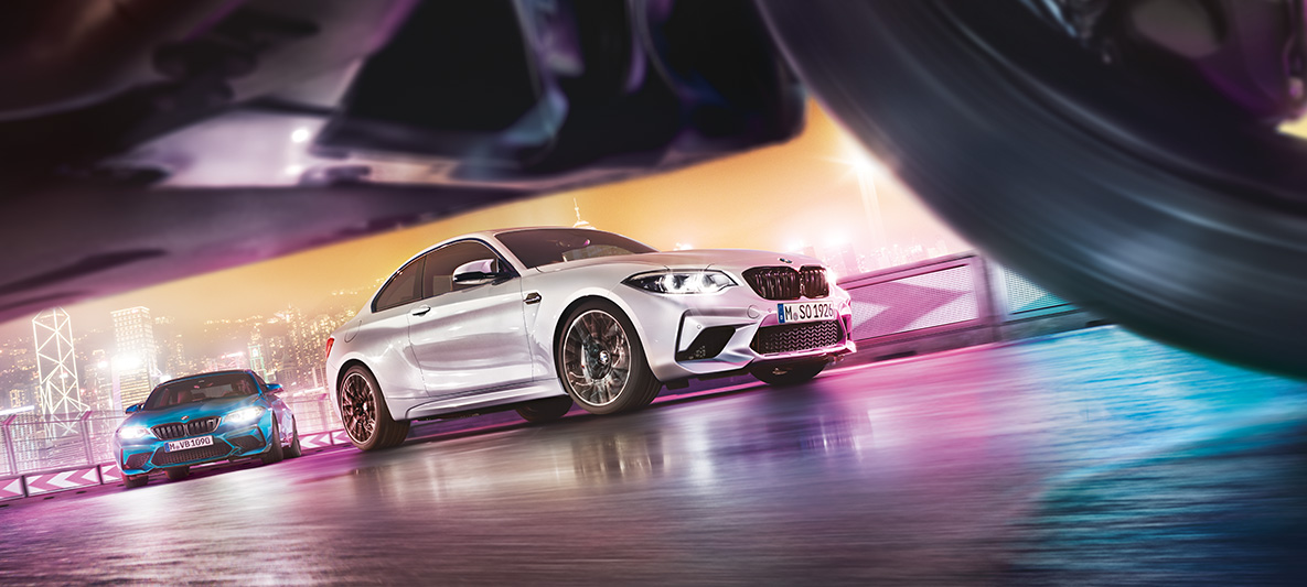 BMW M2 Competition na zrkadliacom sa asfalte Dve BMW M2 Competition na pretekárskej trati pred kulisou nočného mesta z pohľadu úrovne cesty.