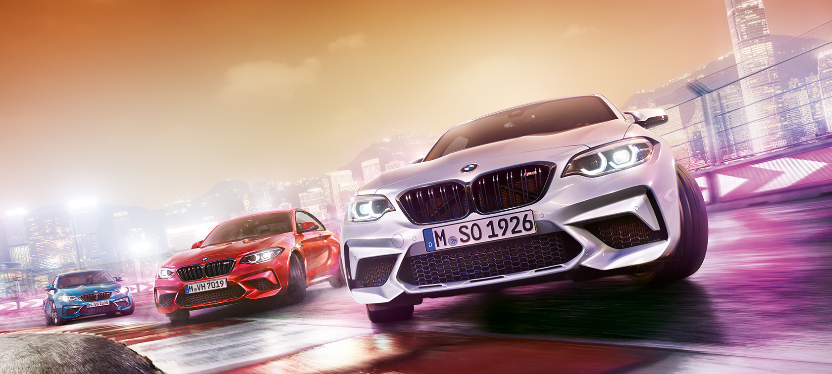 BMW M2 Competition v akcii Tri BMW M2 Competition driftujú na pretekárskej trati pred kulisou nočného mesta.