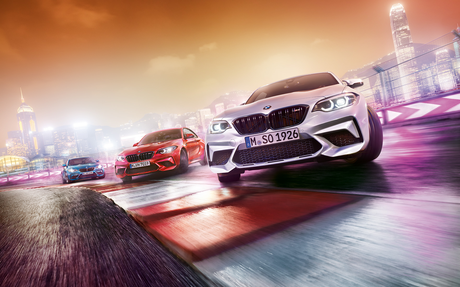 BMW M2 Competition v akcii Tri BMW M2 Competition driftujú na pretekárskej trati pred kulisou nočného mesta.