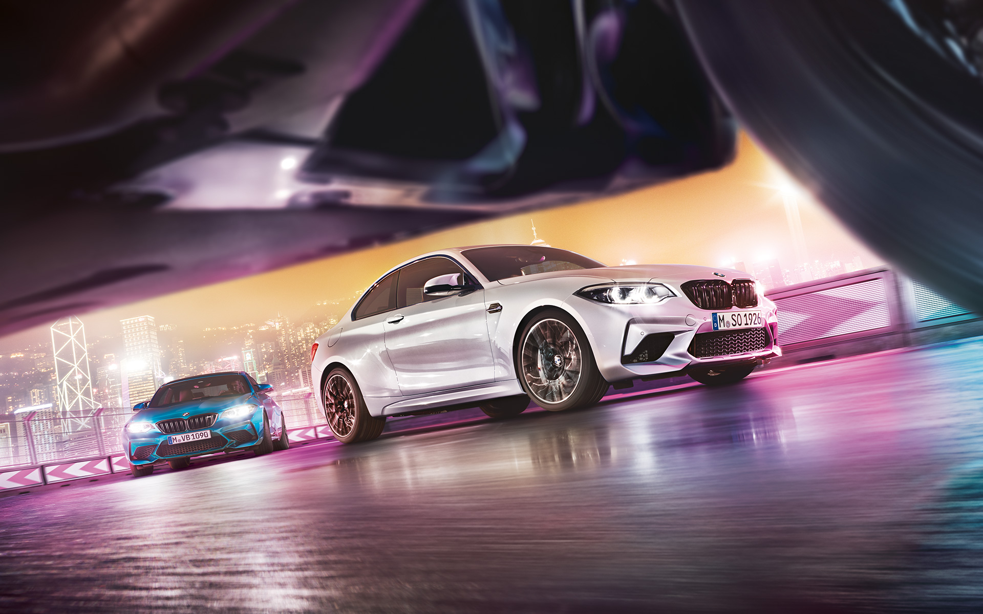 BMW M2 Competition na zrkadliacom sa asfalte Dve BMW M2 Competition na pretekárskej trati pred kulisou nočného mesta z pohľadu úrovne cesty.