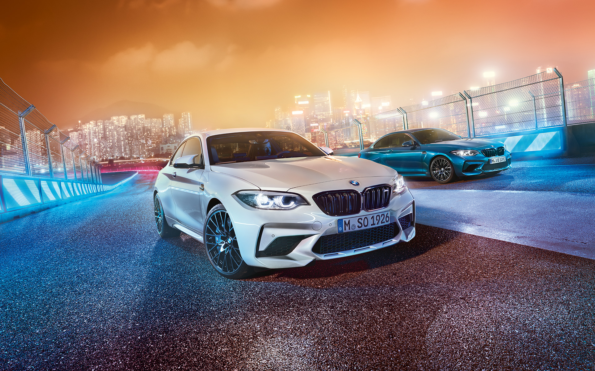 Dve BMW M2 Competition na pretekárskej trati Biele a modré BMW M2 Competition v šikmom prednom pohľade stojace pred kulisou nočného mesta.
