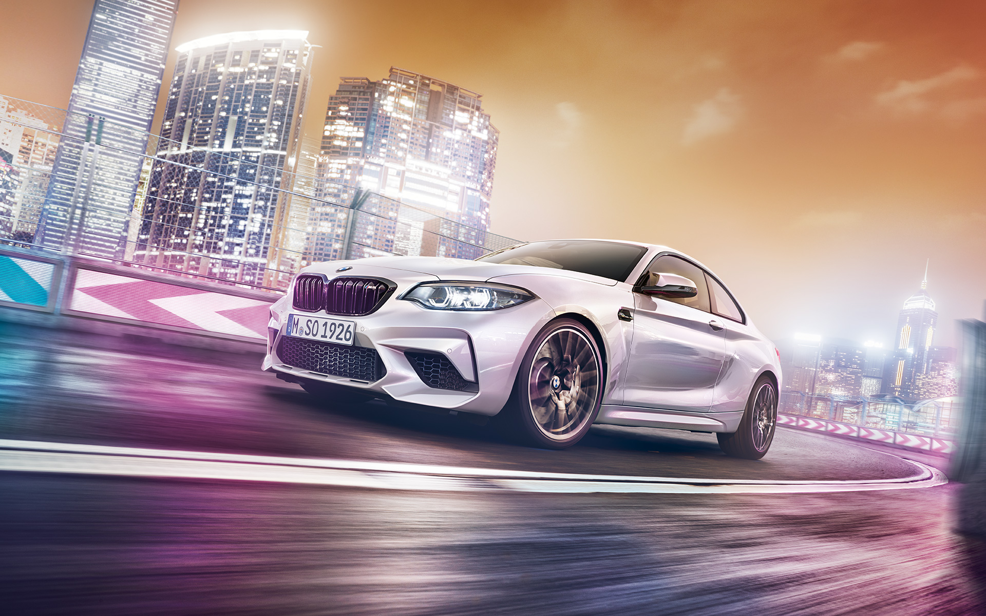 Biele BMW M2 Competition pred nočným mestom Biele BMW M2 Competition jazdí po pretekárskej trati pred kulisou nočného mesta.