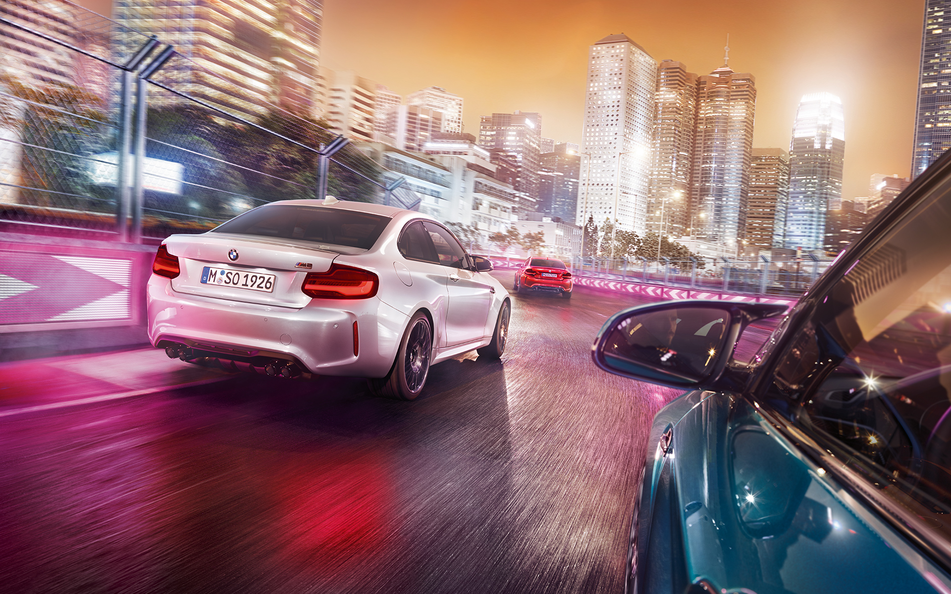 BMW M2 Competition zozadu Tri BMW Competition zozadu jazdia po pretekárskej trati pred osvetlenou kulisou mesta.