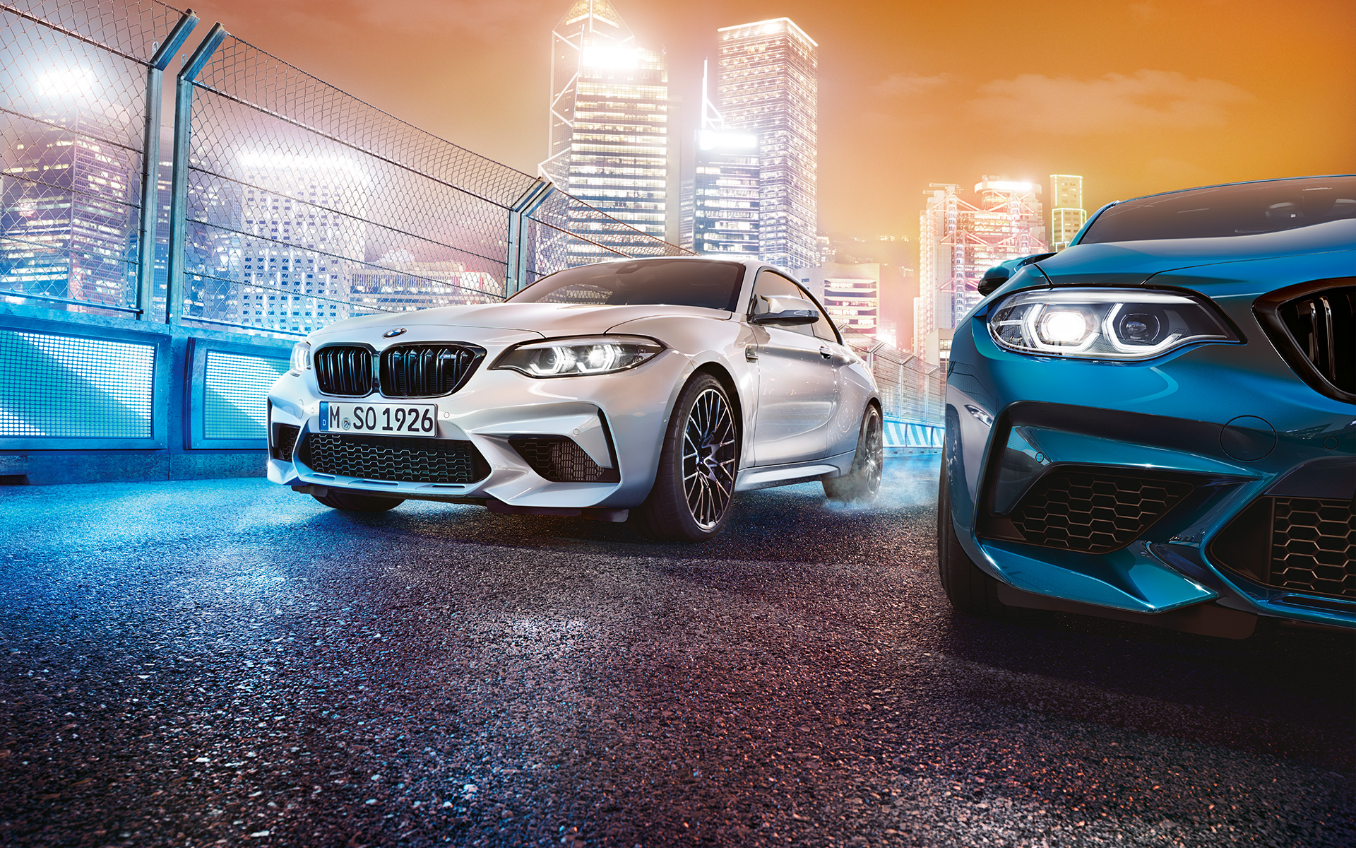 BMW M2 Competition spredu Biele a modré BMW M2 Competition spredu stoja pred kulisou nočného mesta.