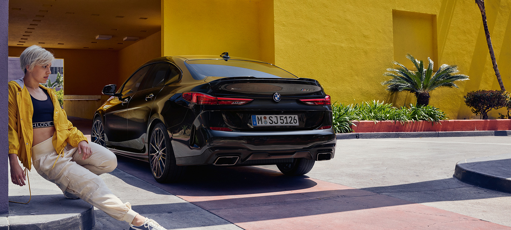 BMW M235i xDrive Gran Coupé trojštvrťový zadný pohľad BMW M235i xDrive Gran Coupé v metalickej farbe Black Sapphire, F44 trojštvrťový zadný pohľad so ženou v popredí