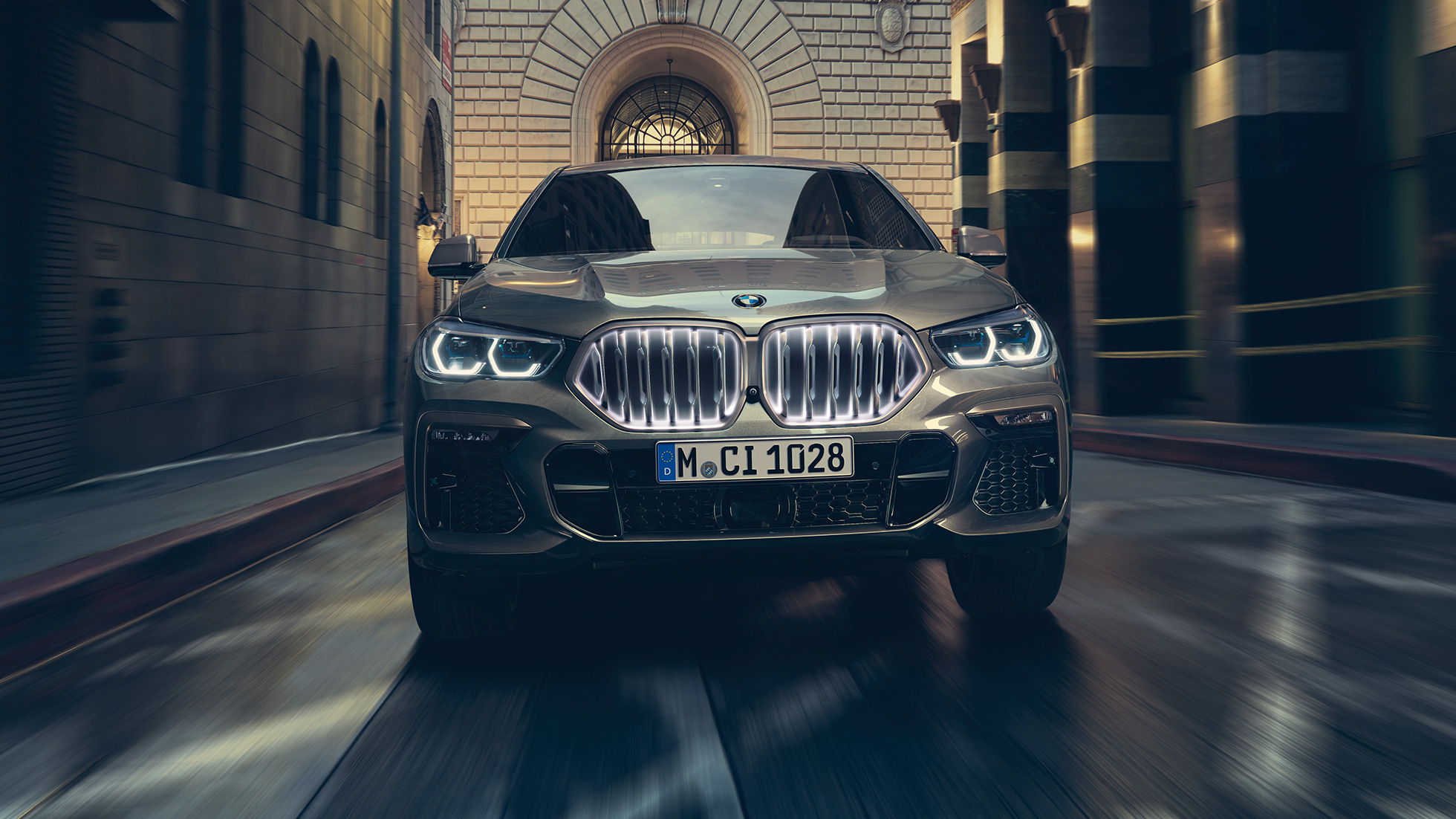 BMW X6 M50i s BMW mriežkou chladiča Iconic Glow. BMW X6 M50i, predný pohľad na vozidlo pred kulisami mesta.