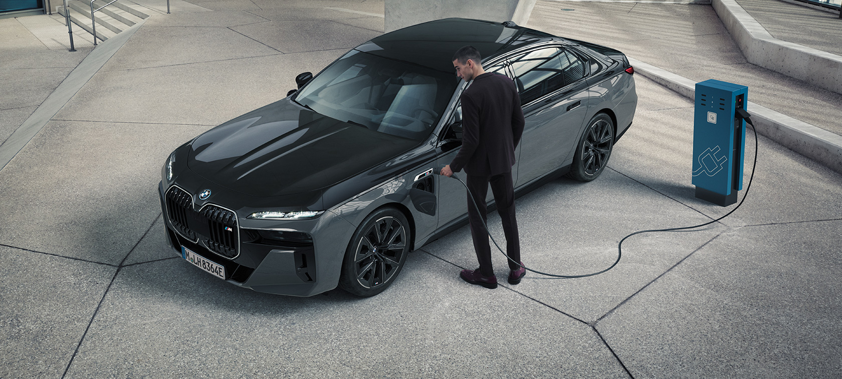 BMW M760e xDrive Sedan G70 M Performance balík BMW Individual dvojfarebná metalíza Black Sapphire Dravite Grey predobočný pohľad muž s nabíjacím káblom pri nabíjacej stanici