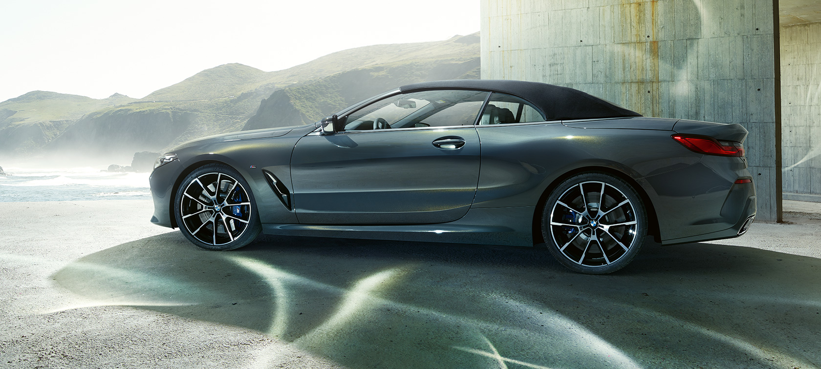 BMW M850i xDrive Cabrio, bočný pohľad. BMW M850i xDrive Cabrio v metalickej farbe Dravit Grey so zatvorenou strechou, bočný pohľad pred horskou scenériou.