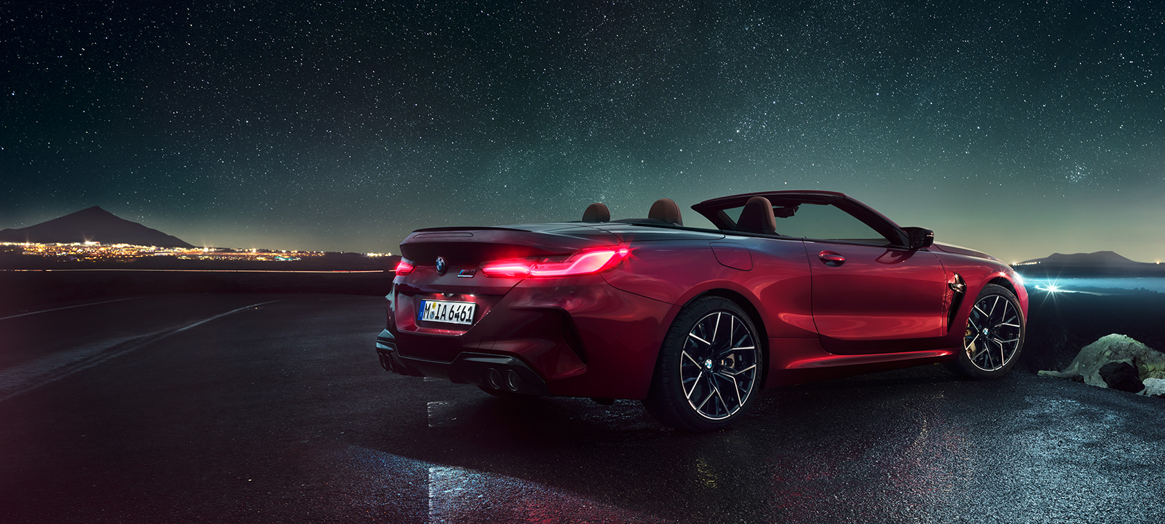 BMW M8 Competition Cabrio, trojštvrťový zadný pohľad. BMW M8 Competition Cabrio v metalickej farbe Motegi Red s otvorenou strechou, trojštvrťový zadný pohľad, vozidlo stojace pod hviezdnou oblohou.