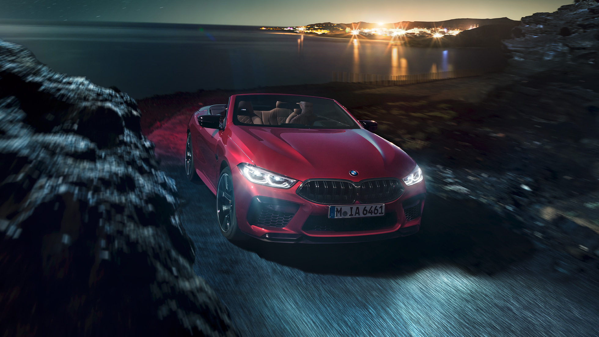 BMW M8 Competition Cabrio, trojštvrťový predný pohľad. BMW M8 Competition Cabrio v metalickej farbe Motegi Red s otvorenou strechou, trojštvrťový bočný pohľad, vozidlo jazdiace v noci po pobrežnej ceste.
