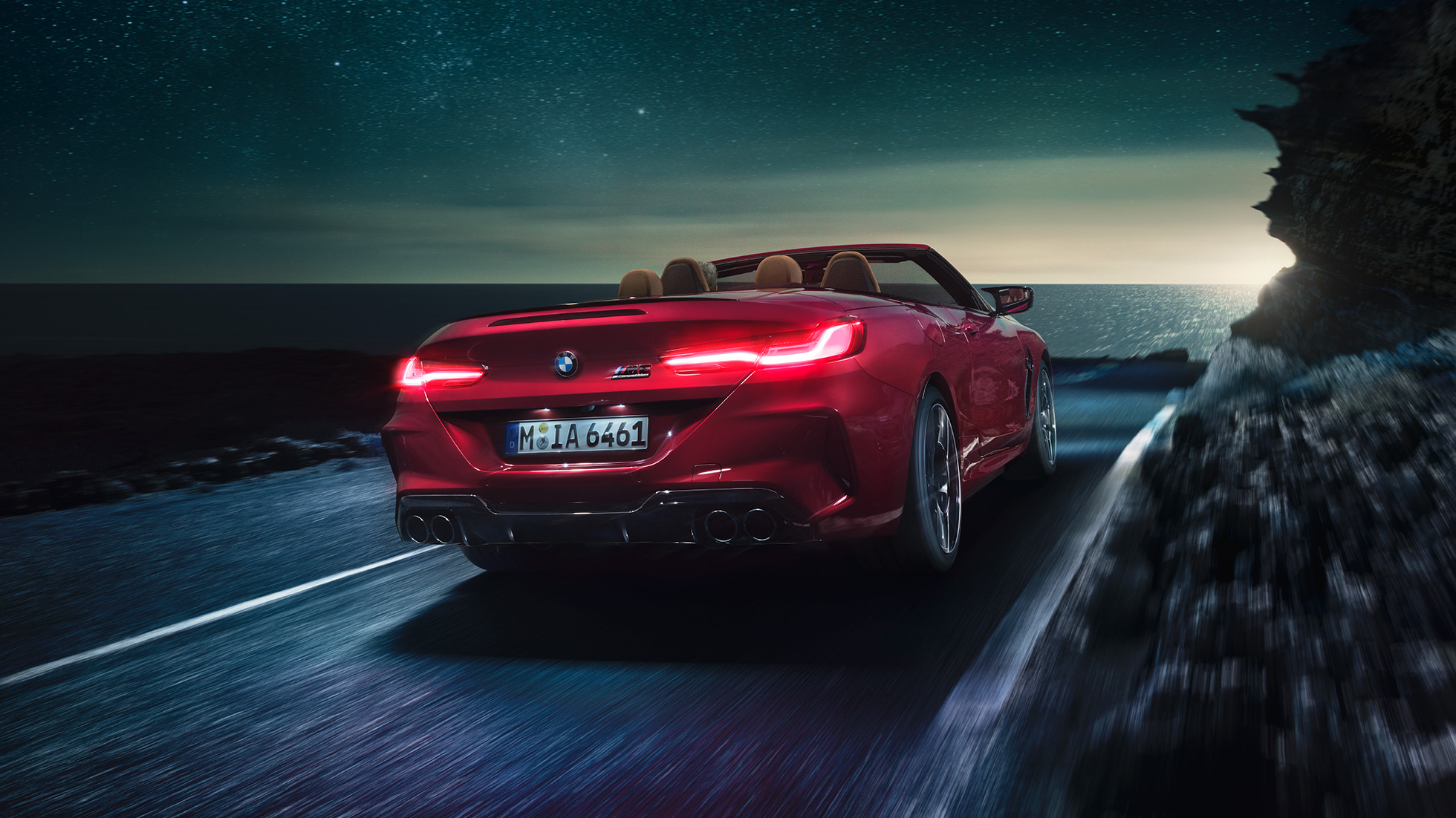 BMW M8 Competition Cabrio, trojštvrťový zadný pohľad. BMW M8 Competition Cabrio v metalickej farbe Motegi Red s otvorenou strechou, trojštvrťový zadný pohľad, vozidlo jazdiace v noci po pobrežnej ceste.