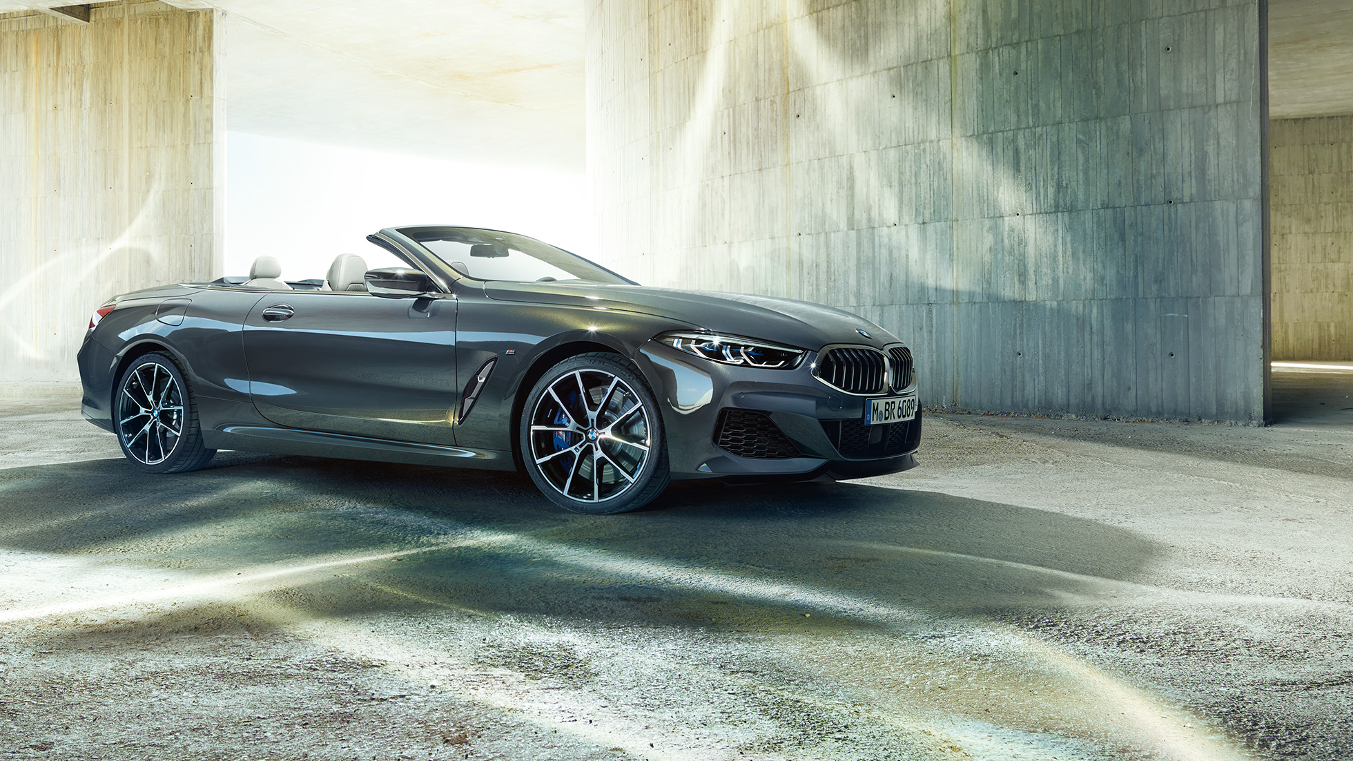 BMW M850i xDrive Cabrio, trojštvrťový bočný pohľad. BMW M850i xDrive Cabrio v metalickej farbe Dravit Grey s otvorenou strechou, trojštvrťový bočný pohľad, vozidlo stojace pred betónovou budovou.