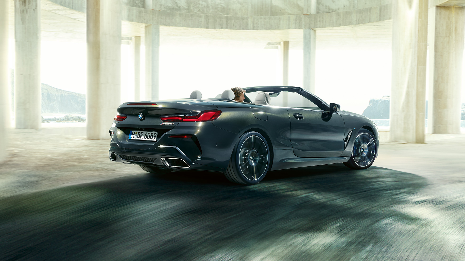 BMW M850i xDrive Cabrio, trojštvrťový zadný pohľad. BMW M850i xDrive Cabrio v metalickej farbe Dravit Grey s otvorenou strechou, trojštvrťový zadný pohľad, vozidlo jazdiace cez betónovú budovu.
