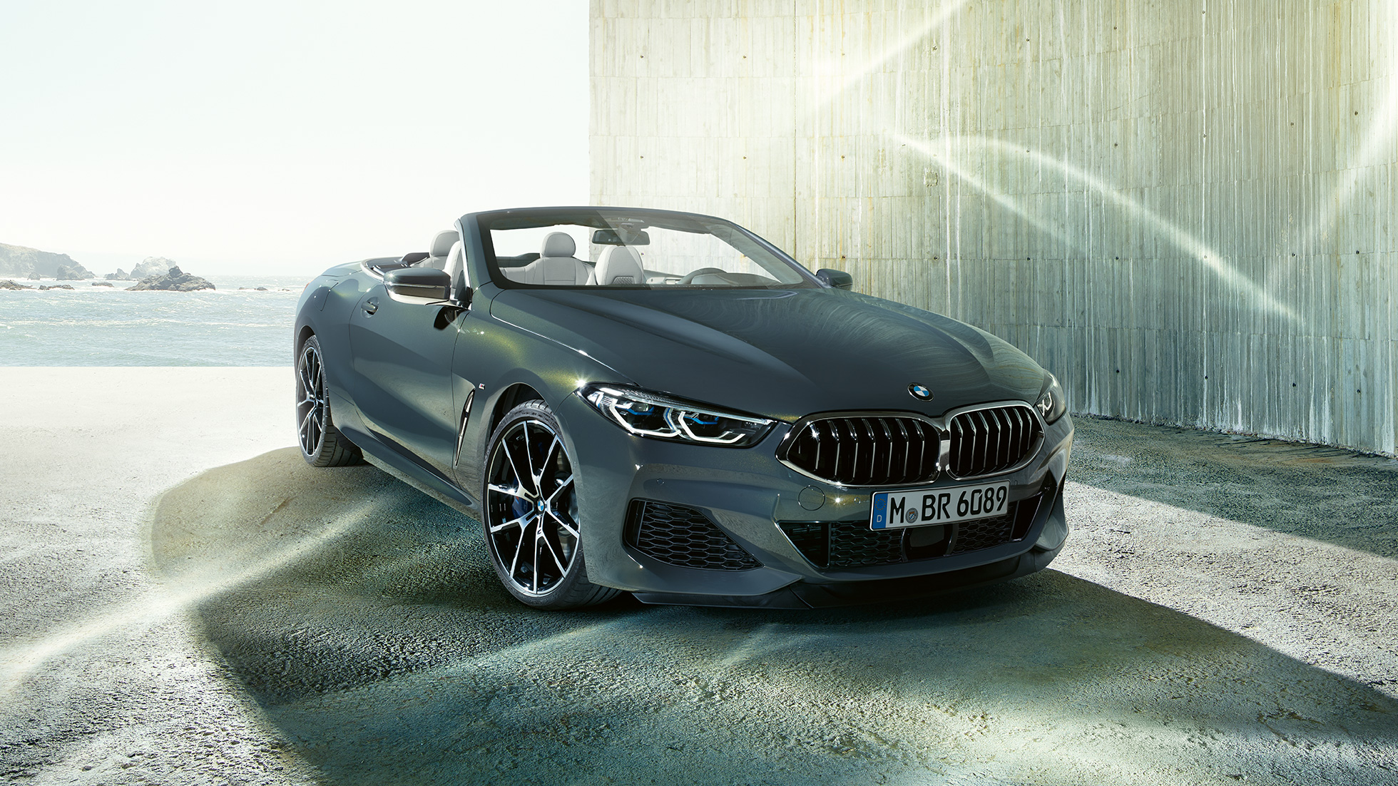 BMW M850i xDrive Cabrio, trojštvrťový predný pohľad. BMW M850i xDrive Cabrio v metalickej farbe Dravit Grey s otvorenou strechou, trojštvrťový predný pohľad, vozidlo stojace na pobreží.