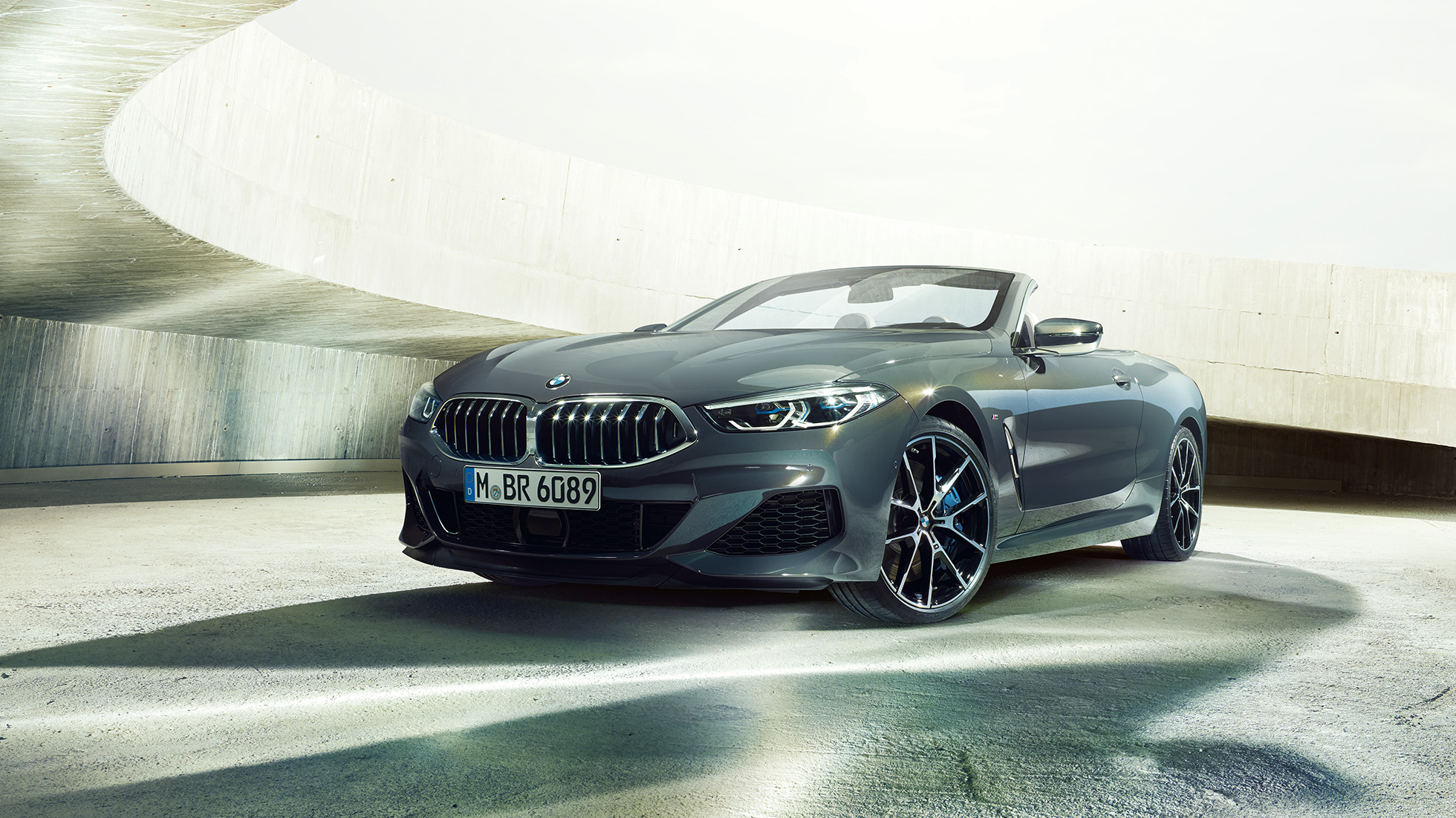 BMW M850i xDrive Cabrio, trojštvrťový predný pohľad. BMW M850i xDrive Cabrio v metalickej farbe Dravit Grey s otvorenou strechou, trojštvrťový predný pohľad, vozidlo stojace v betónovej budove.