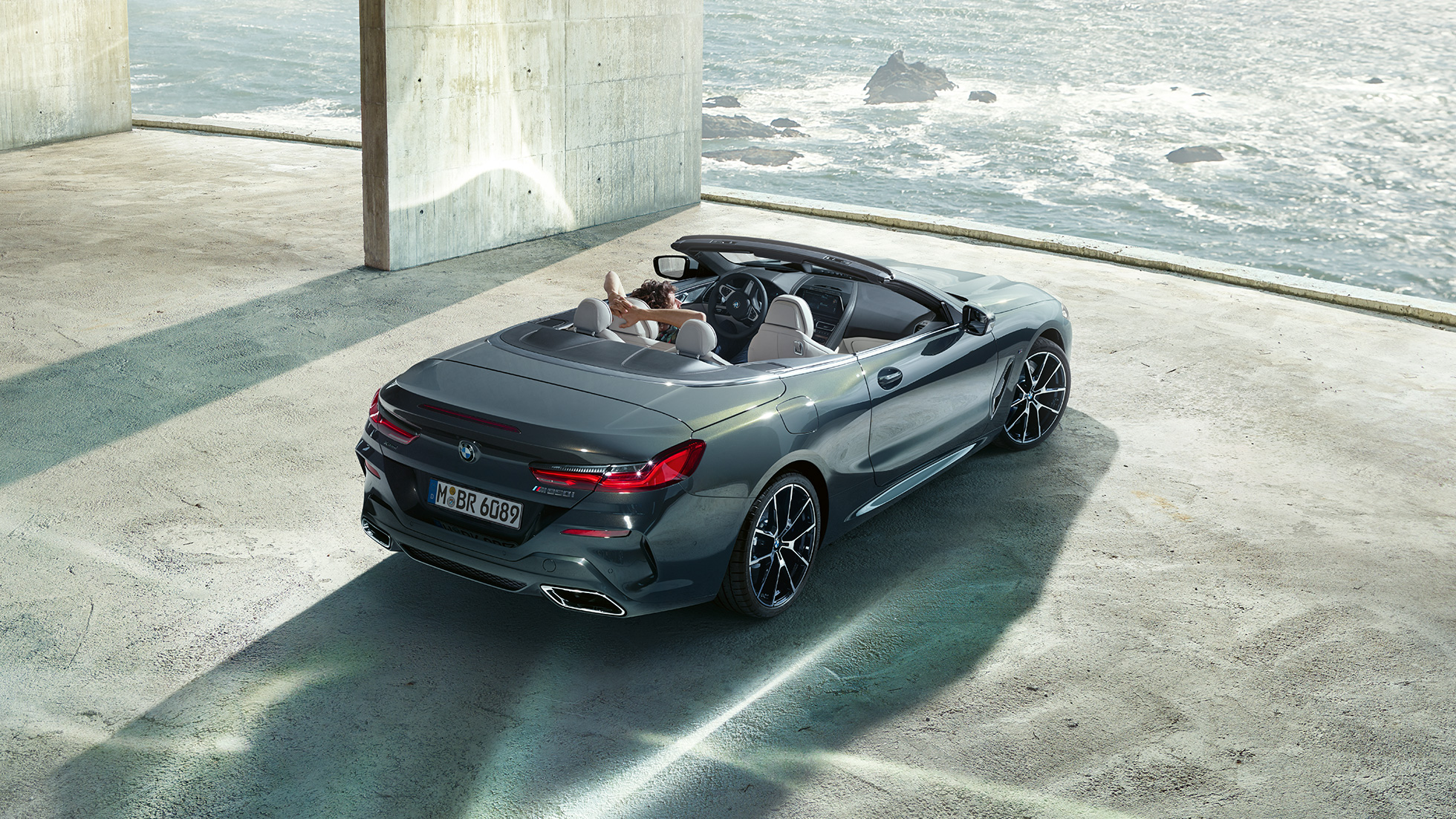 BMW M850i xDrive Cabrio, pohľad z vtáčej perspektívy. BMW M850i xDrive Cabrio v metalickej farbe Dravit Grey s otvorenou strechou, pohľad z vtáčej perspektívy, vozidlo odparkované v betónovej budove na brehu oceánu.