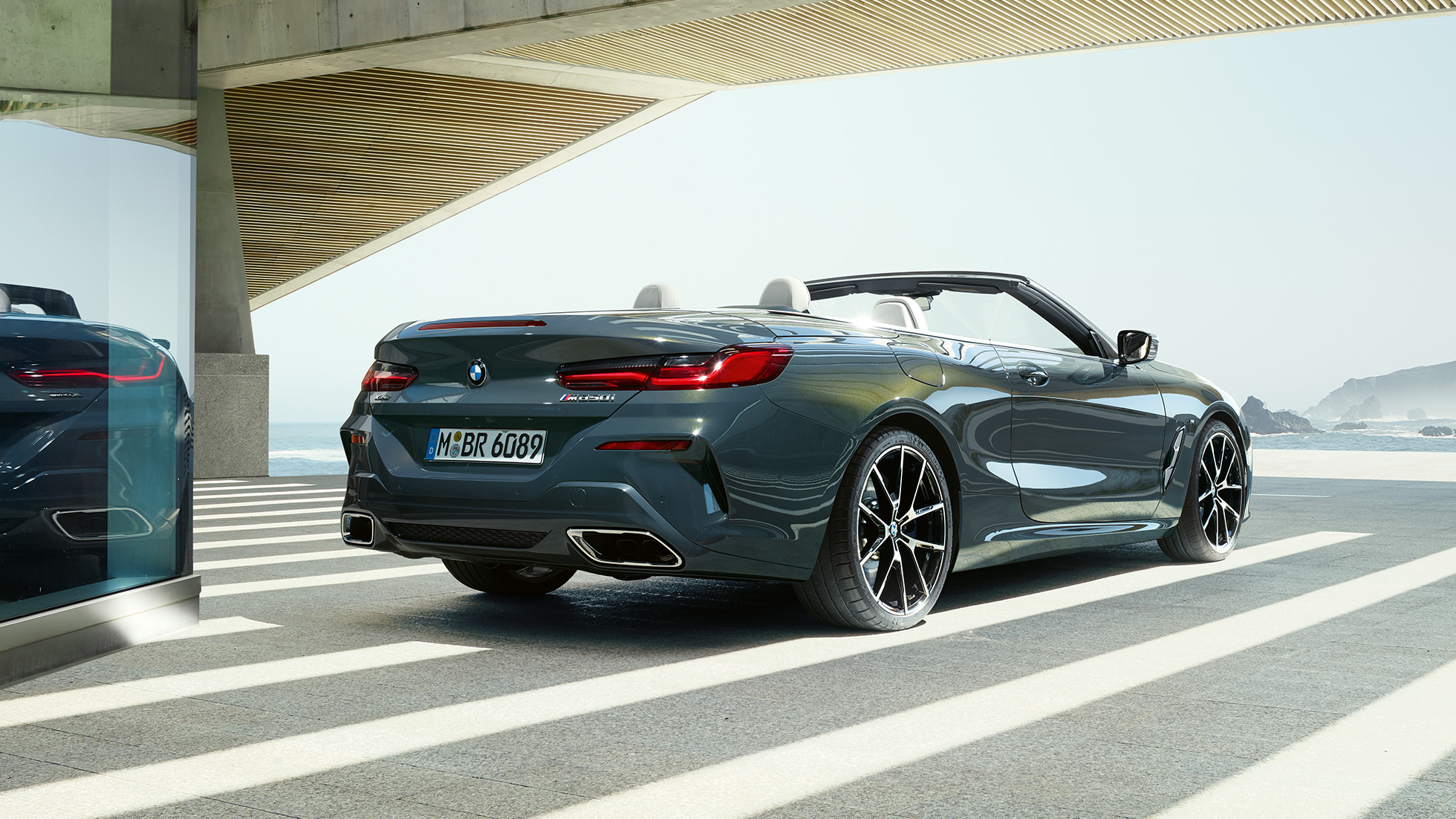 BMW M850i xDrive Cabrio, trojštvrťový zadný pohľad. BMW M850i xDrive Cabrio v metalickej farbe Dravit Grey s otvorenou strechou, trojštvrťový zadný pohľad, stojace vozidlo s výhľadom na oceán.