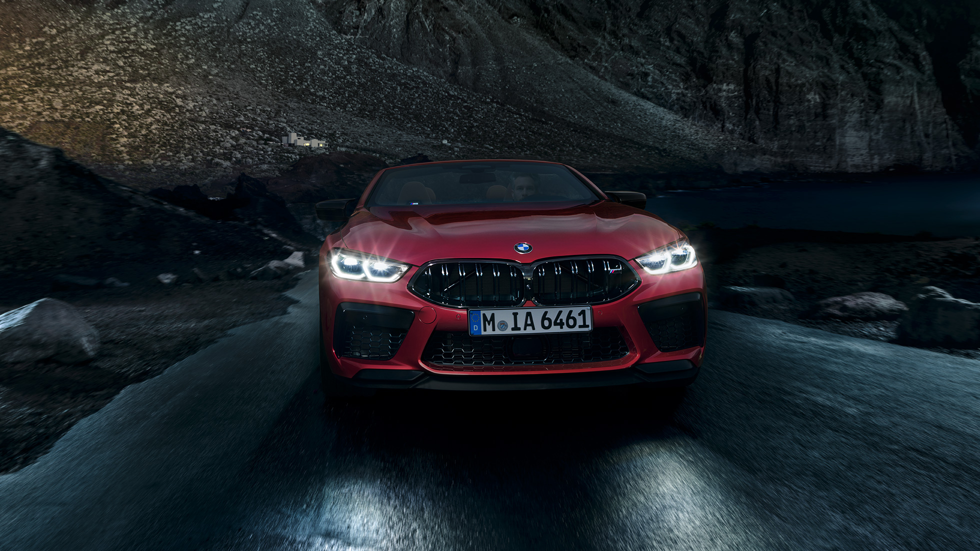 BMW M8 Competition Cabrio, predný pohľad. BMW M8 Competition Cabrio v metalickej farbe Motegi Red s rozsvietenými svetlami, predný pohľad, vozidlo jazdiace v noci.