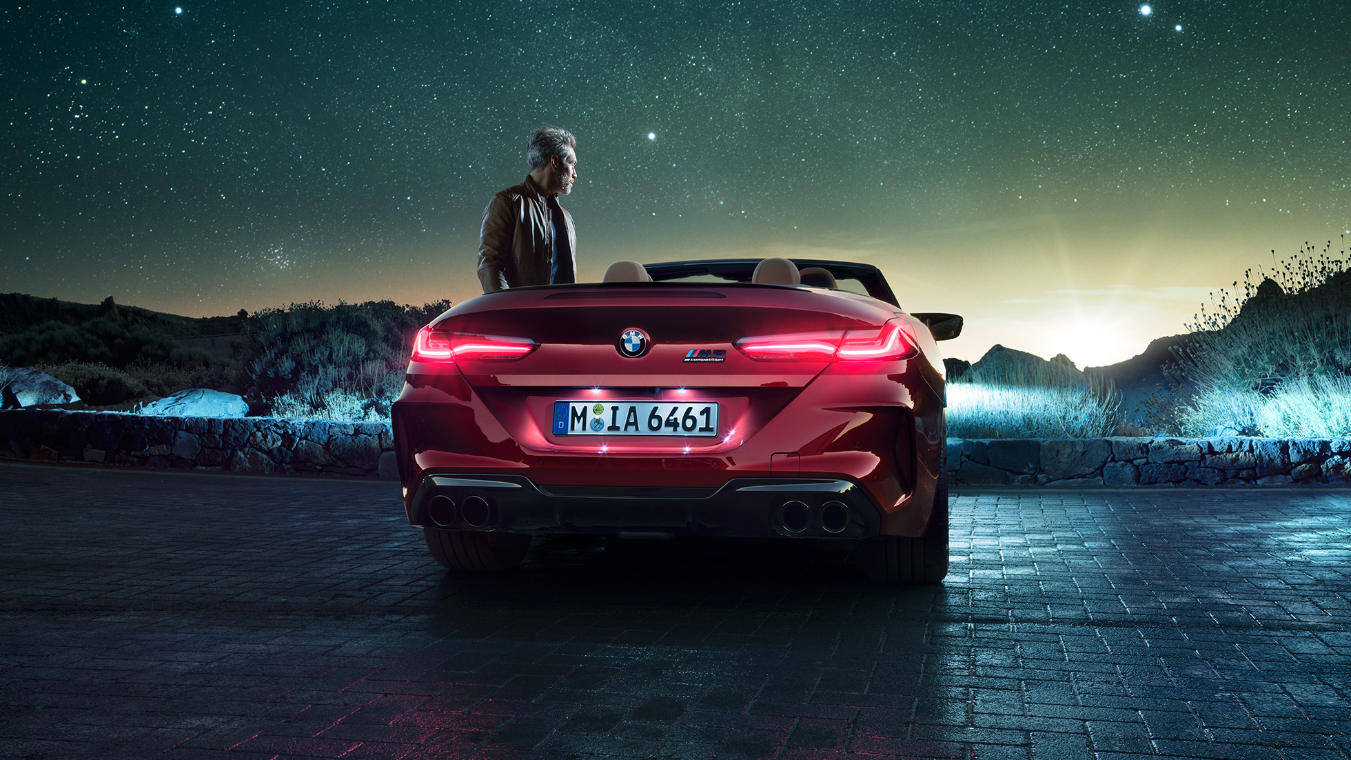 BMW M8 Competition Cabrio, zadný pohľad. BMW M8 Competition Cabrio v metalickej farbe Motegi Red s otvorenou strechou, zadný pohľad, stojace vozidlo s mužom pod hviezdnou oblohou.