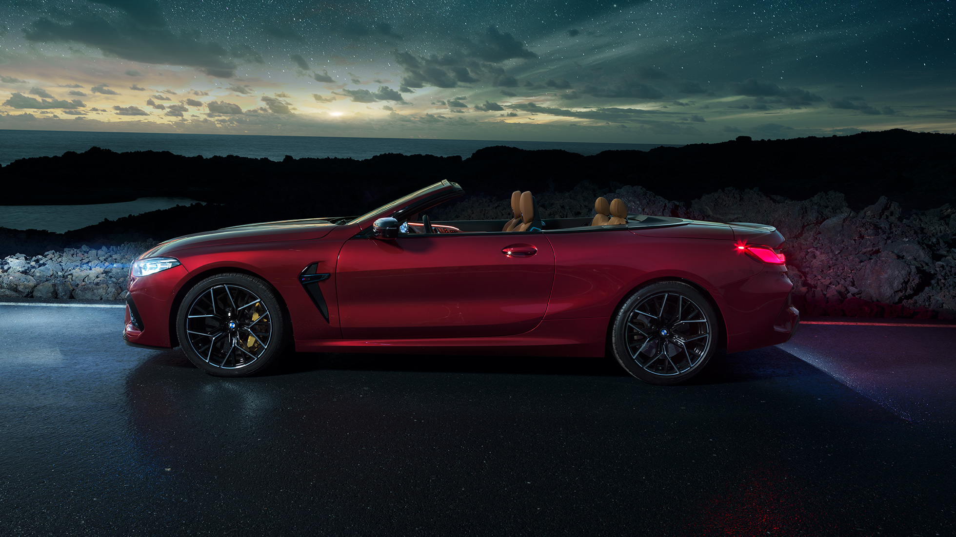 BMW M8 Competition Cabrio, bočný pohľad. BMW M8 Competition Cabrio v metalickej farbe Motegi Red s otvorenou strechou, bočný pohľad, vozidlo stojace v noci pod hviezdnou oblohou.