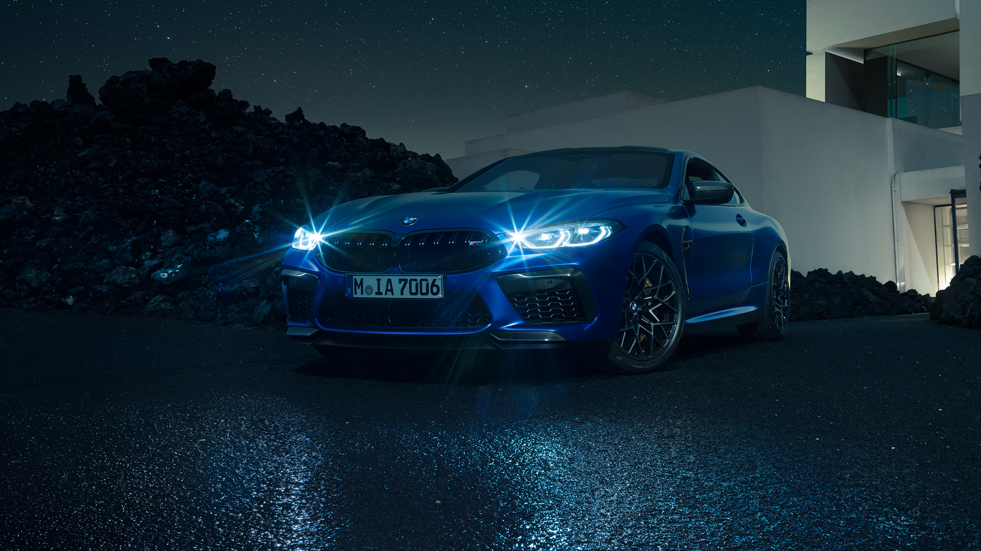 BMW M8 Competition Coupé, trojštvrťový predný pohľad. BMW M8 Competition Coupé v metalickej farbe Marina Bay Blue so žiarivými LED svetlometmi, statický trojštvrťový predný pohľad pred priečelím vily počas noci.