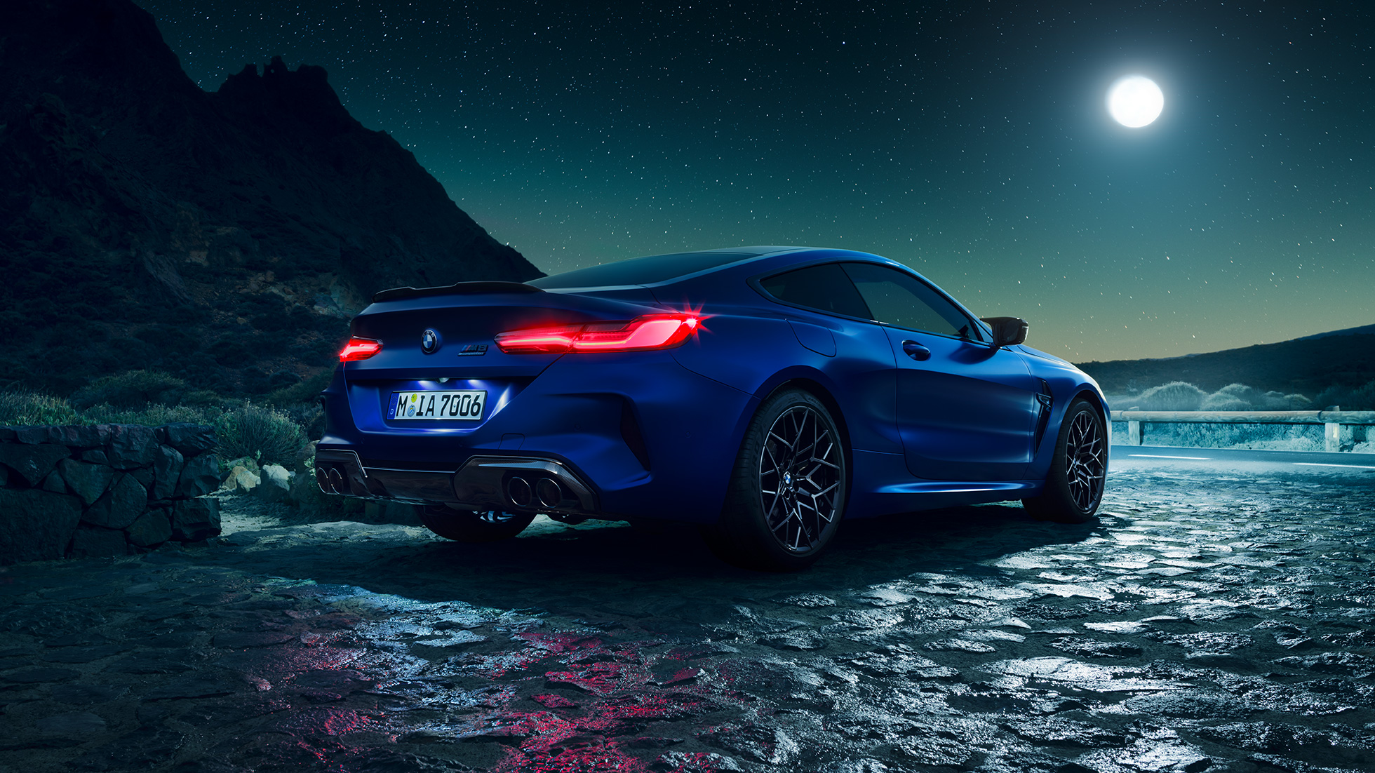 BMW M8 Competition Coupé, trojštvrťový zadný pohľad. BMW M8 Competition Coupé v metalickej farbe Marina Bay Blue s rozsvietenými LED zadnými svetlami, trojštvrťový statický zadný pohľad počas splnu mesiaca.