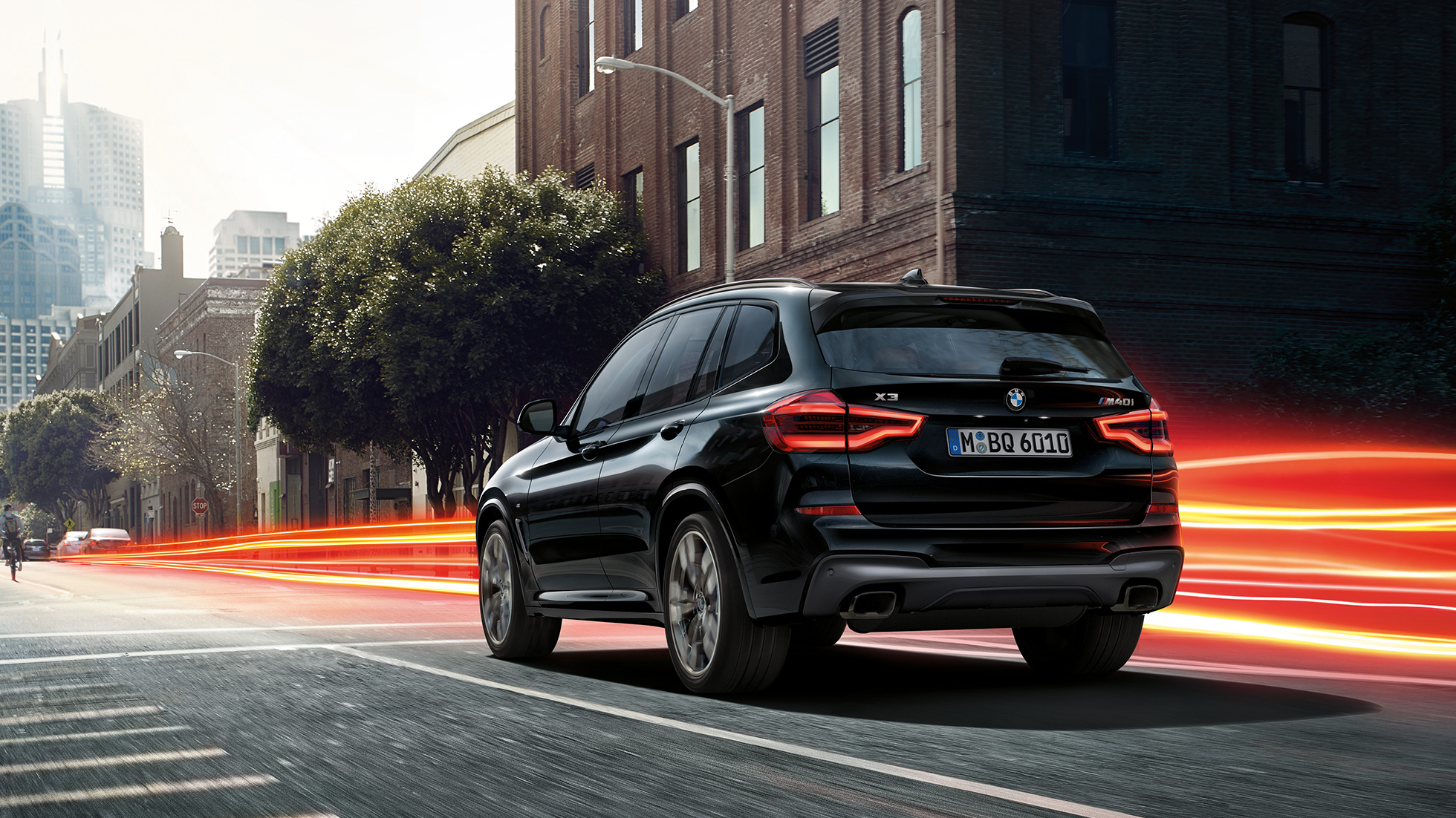 BMW X3 M40i, trojštvrťový zadný pohľad. BMW X3 M40i v metalickej farbe Black Sapphire, jazda, exteriér, trojštvrťový zadný pohľad pred kulisami mesta so svetelnými pásmi.