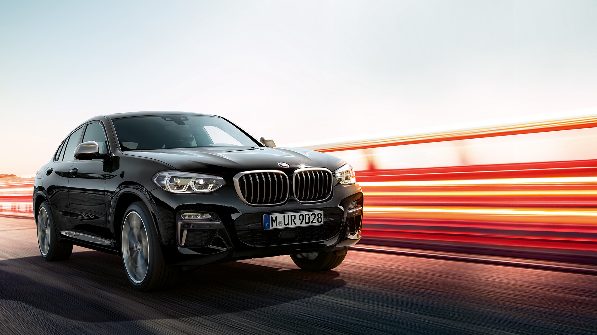 BMW X4 M40i, trojštvrťový zadný pohľad. BMW X4 M40i v metalickej farbe Black Sapphire, jazda, exteriér, trojštvrťový zadný pohľad so svetelnými pásmi.