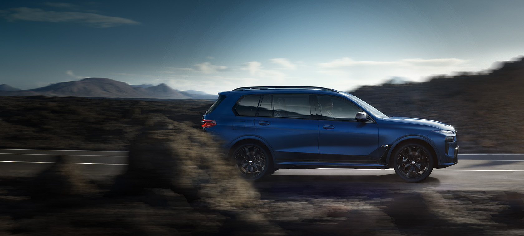 BMW X7 M60i xDrive G07 LCI Frozen Marina Bay Blue bočný pohľad jazdný na ceste BMW X7 M60i xDrive G07 LCI Frozen Marina Bay Blue bočný pohľad jazdný na ceste s horami v pozadí