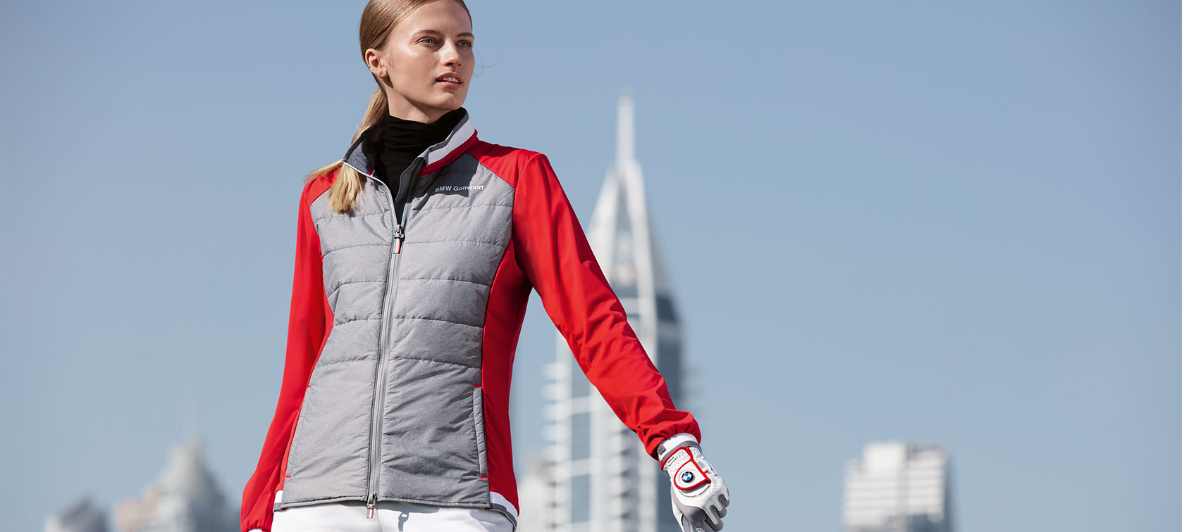 BMW Golfsport dámska bunda. Mladá žena na obrázku prezentuje prémiové produkty z BMW Golfsport kolekcie na pozadí panorámy veľkomesta.