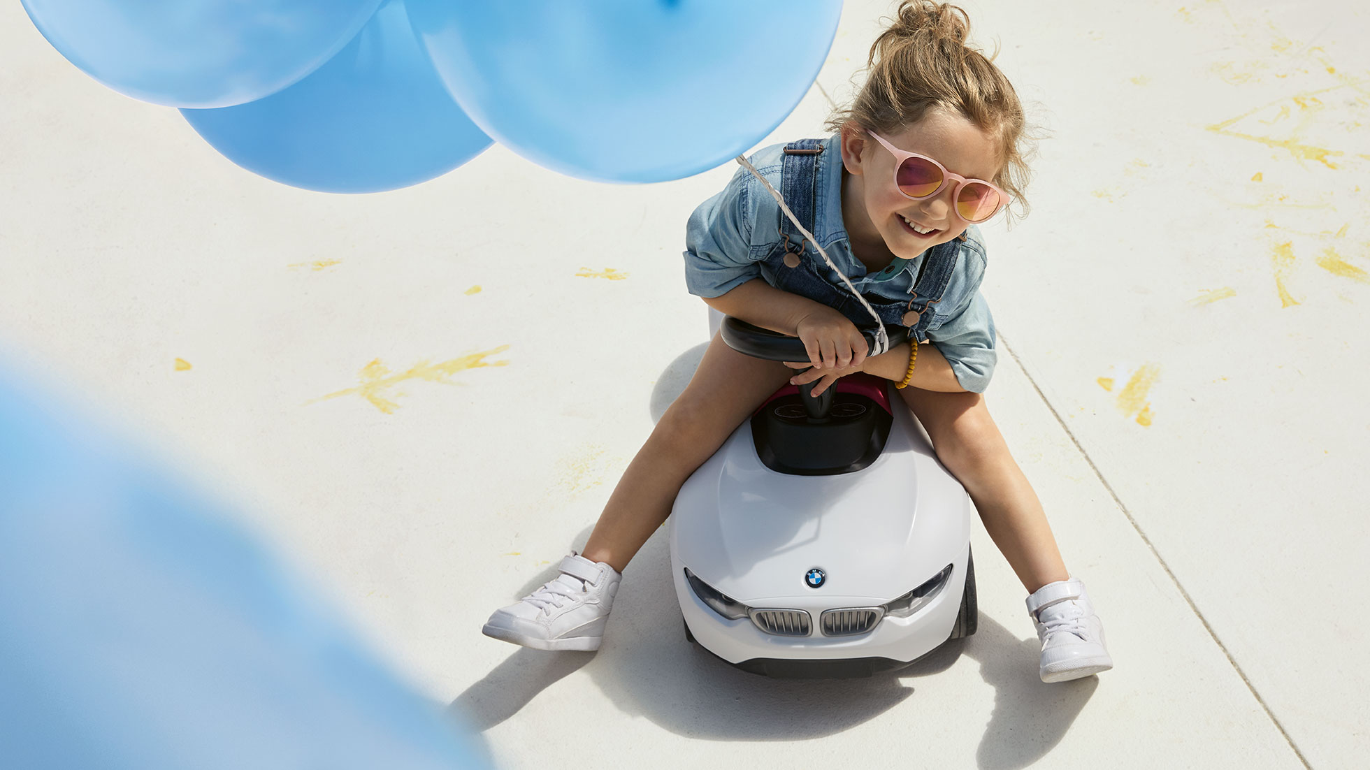 BMW Detská kolekcia. Dievča s ružovými slnečnými okuliarmi a modrým balónom sedí šťastne na BMW Baby Racer.